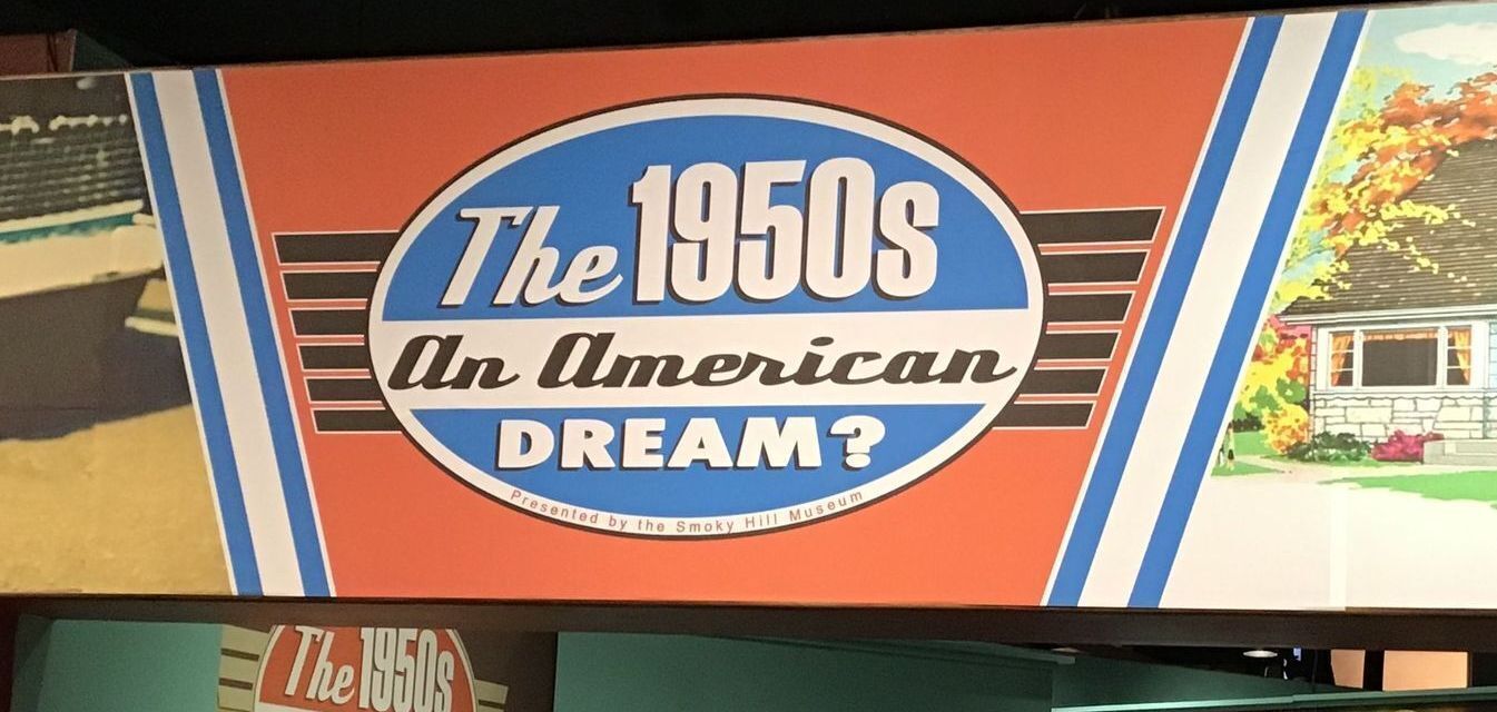 sign at the Smoky Hill Museum showing a front end of a 1950s car and a 1950s home with the exhibit title, The 1950s: An American Dream?"