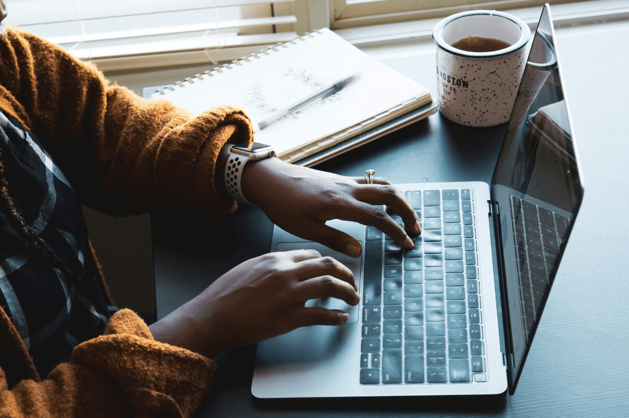 Photo of hands typing on a laptop next to a notebook and cup of coffee