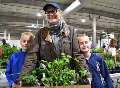 A mom wearing a baseball cap holds a flat of plants with her two twin boys in blue shirts standing on either side of her. 
