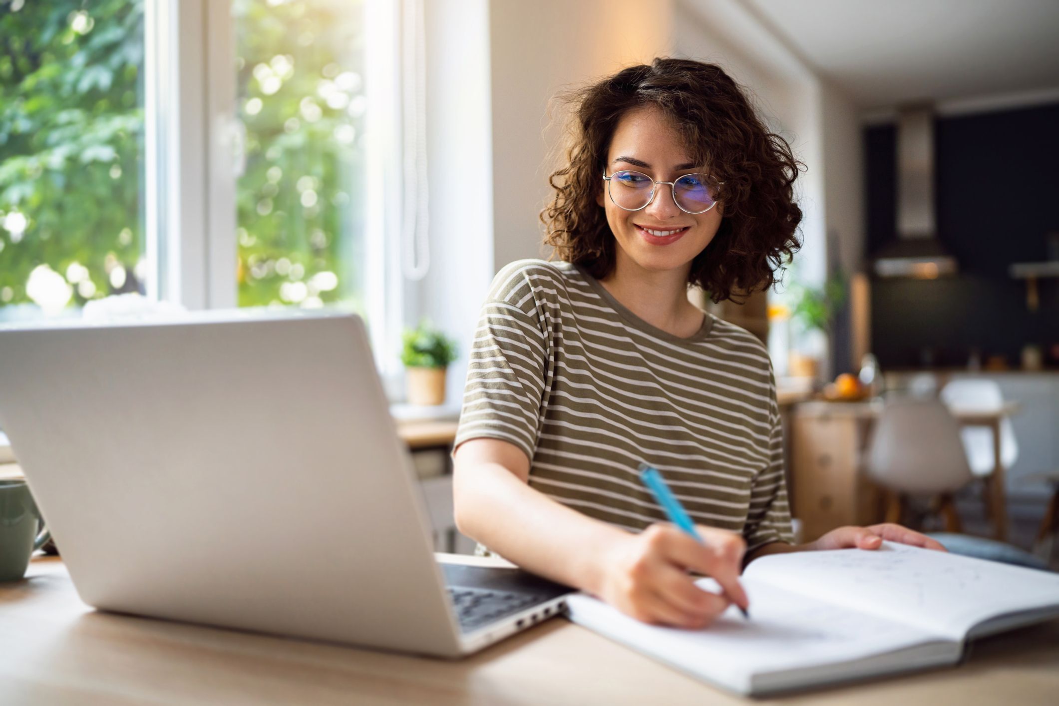 Image Description: an smiling woman looking at an open laptop and taking notes in a notebook