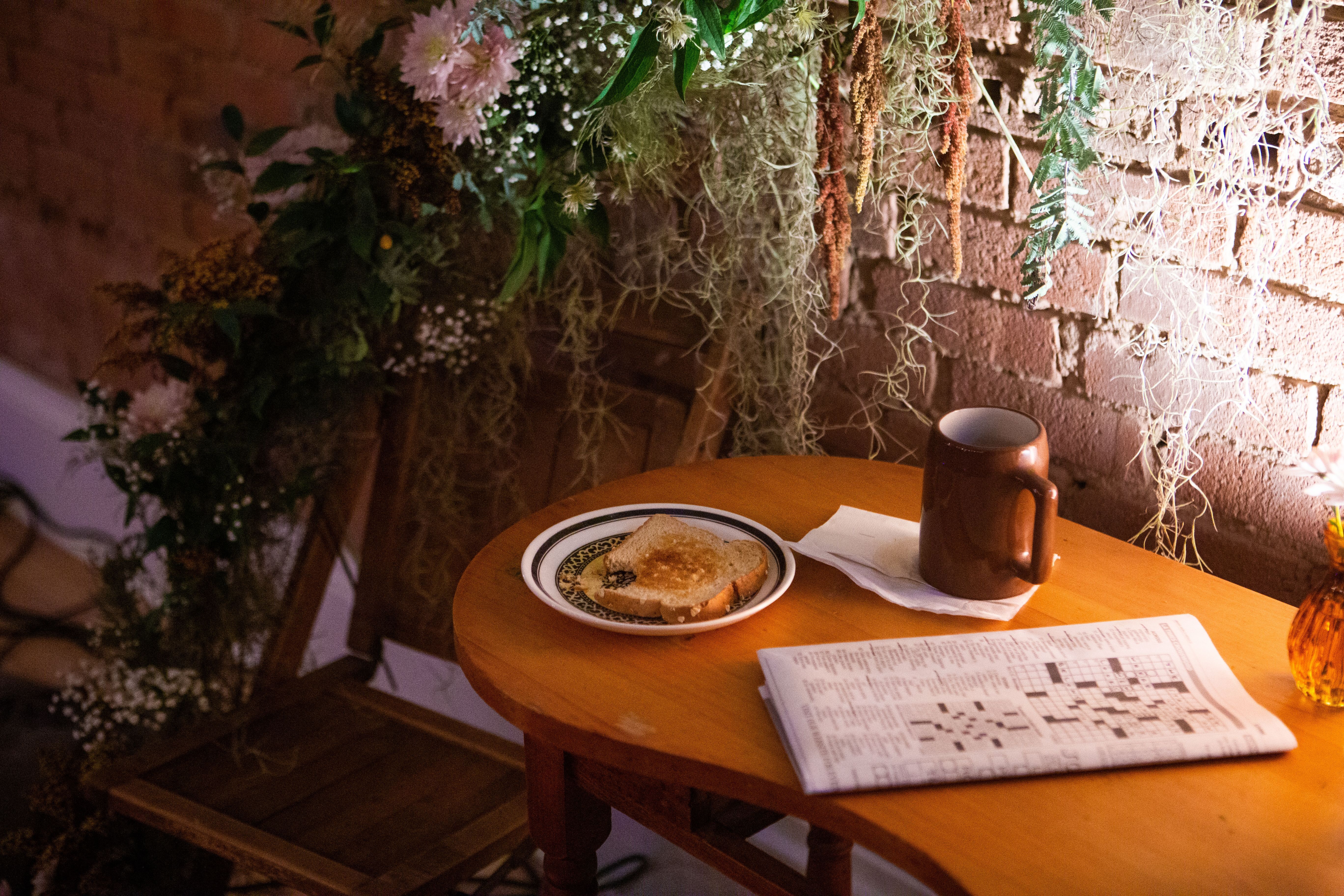 A floral installation frames the background of a table, with a plate of toast, a crossword puzzle, and a mug. 