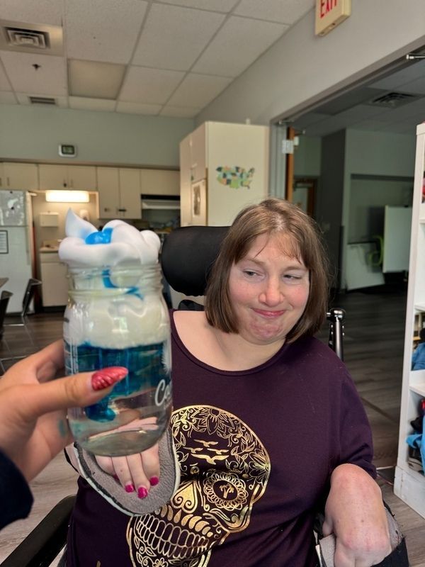 A women in a wheelchair smiling next a staff's hand that this holding up a science project they did. the science project is a mason jar with shaving cream, water, and blue dye to show how clouds make rain. 