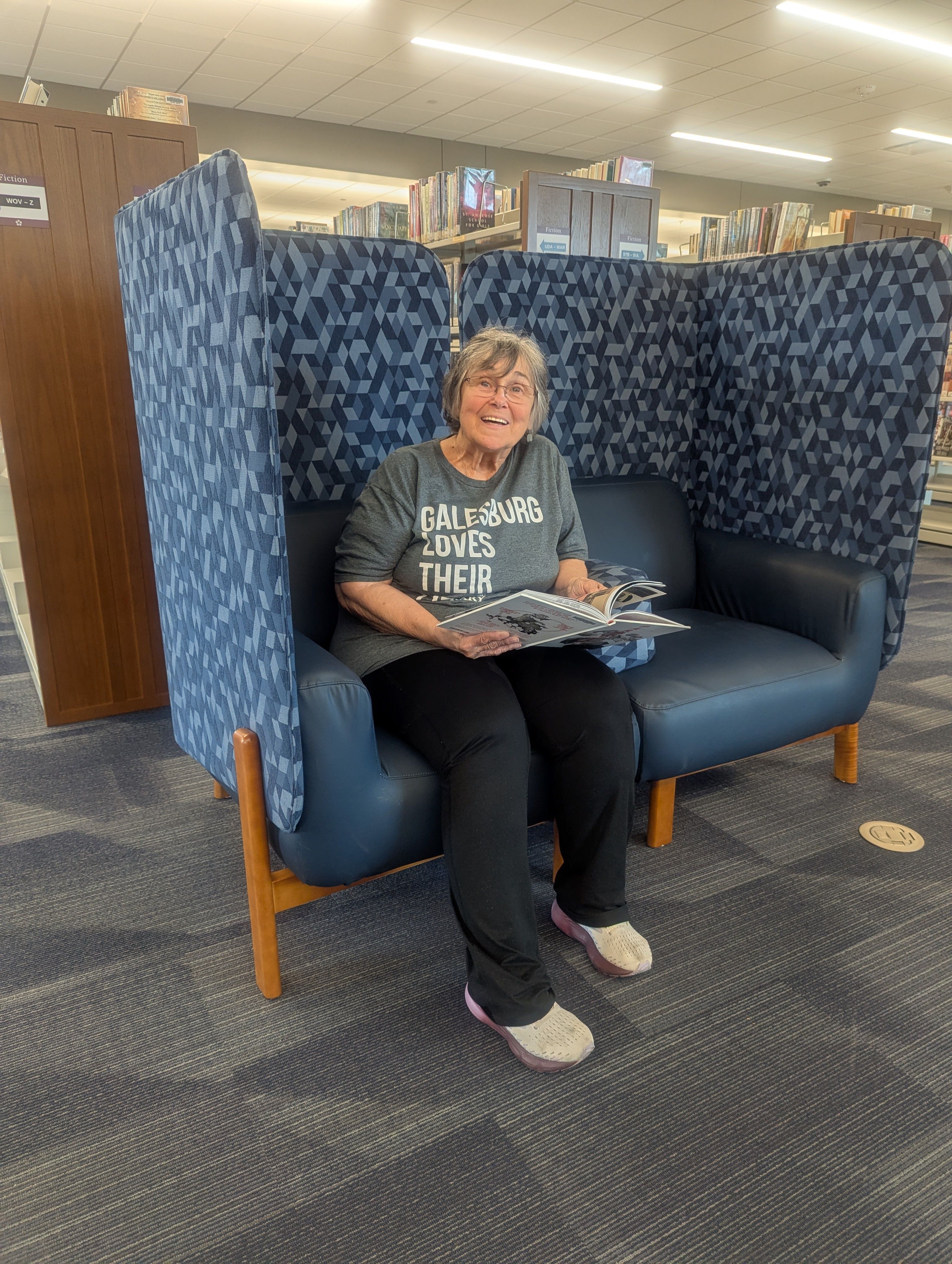 An older woman with short gray hair and glasses smiles while seated in a blue cushioned privacy chair at the Galesburg Public Library. She is holding an open magazine and wearing a shirt that reads “Galesburg Loves Their Library.” Shelves of books and a c