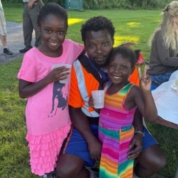 Kalmoi wears a bright orange shirt and is crouching with his two young daughters, both in bright dresses and holding cups of juice.