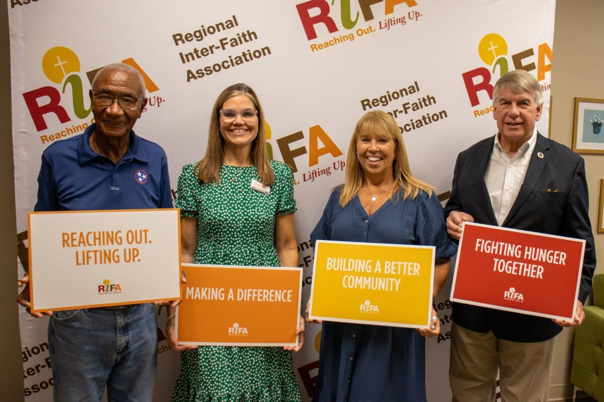 Four people holding signs with messages about community and hunger relief in front of a Regional Inter-Faith Association backdrop.