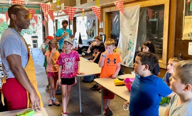 Chef Anwar, dressed in colorful red shorts and a yellow apron teaches a group of children on the right about cooking with nutritious foods