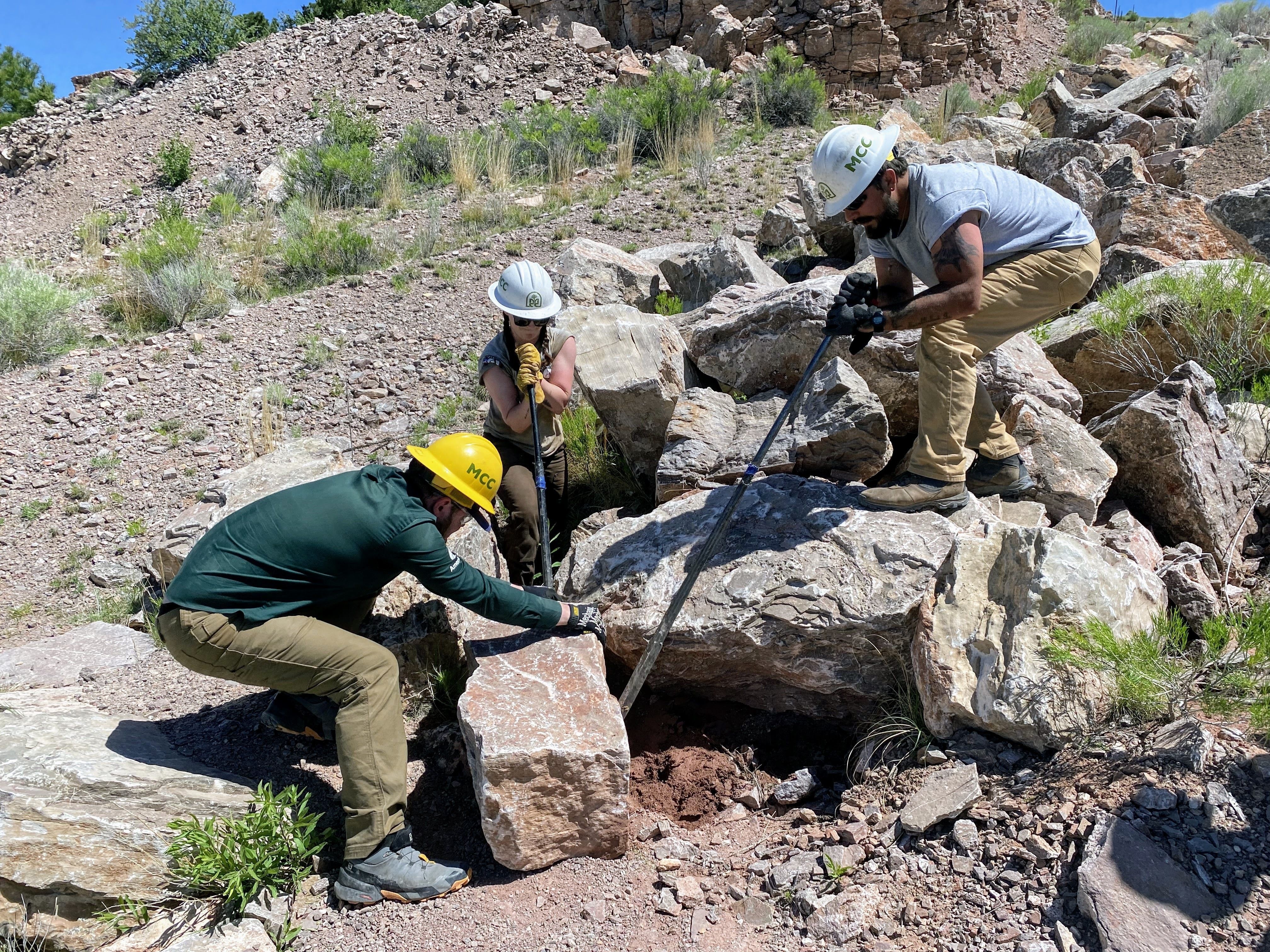 Three people are clustered around a rock, working to move it. Two people on the back and the side are using rock bars to leverage against it, and the third person is using their gloved hands to pull it away.