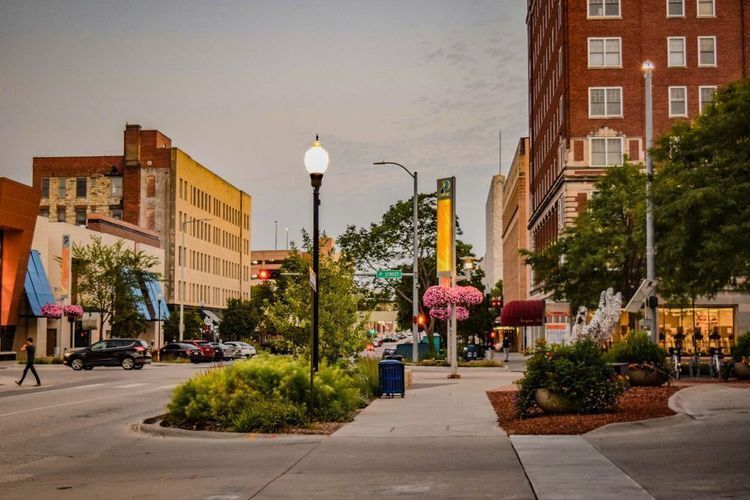 Twilight view of downtown Lincoln, Nebraska at the P Street District intersection, featuring a glowing globe street lamp, landscaped median with ornamental grasses, pink flowering trees, and historic brick buildings lining both sides of the street.