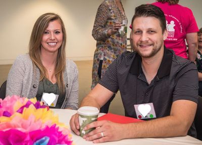 A woman and man sit side-by-side at a conference table smiling at the camera. Some conference goers can be seen in the backgroud.