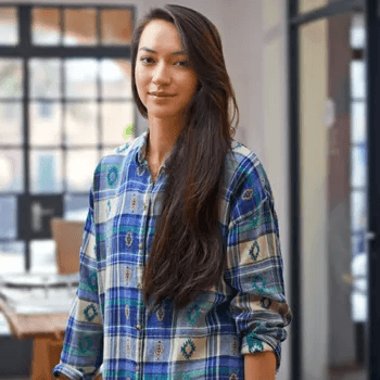 A woman with long straight dark hair is standing indoors wearing a plaid button-down shirt in blue and green tones. She is in a modern space with large grid-pattern windows and part of a table with documents visible in the background.