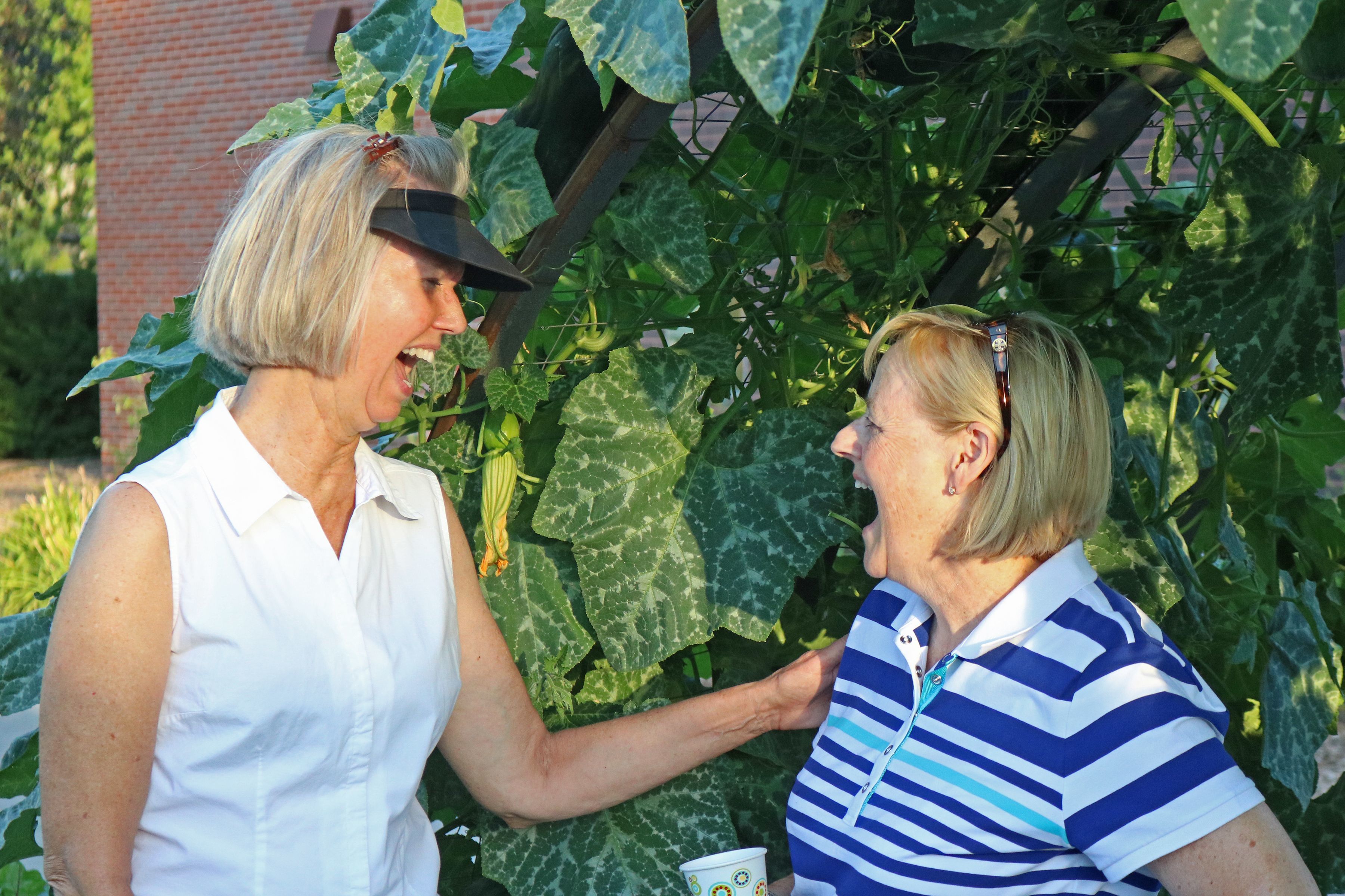 Two female gardeners talking and laughing together in front of lush, green shrubbery. 