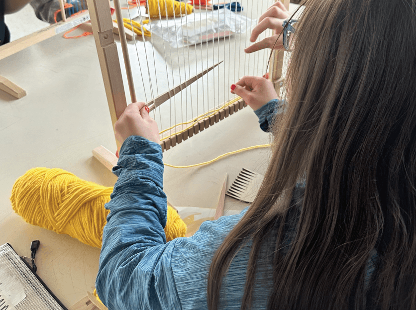 Photo of a student weaving yellow yarn onto a loom.
