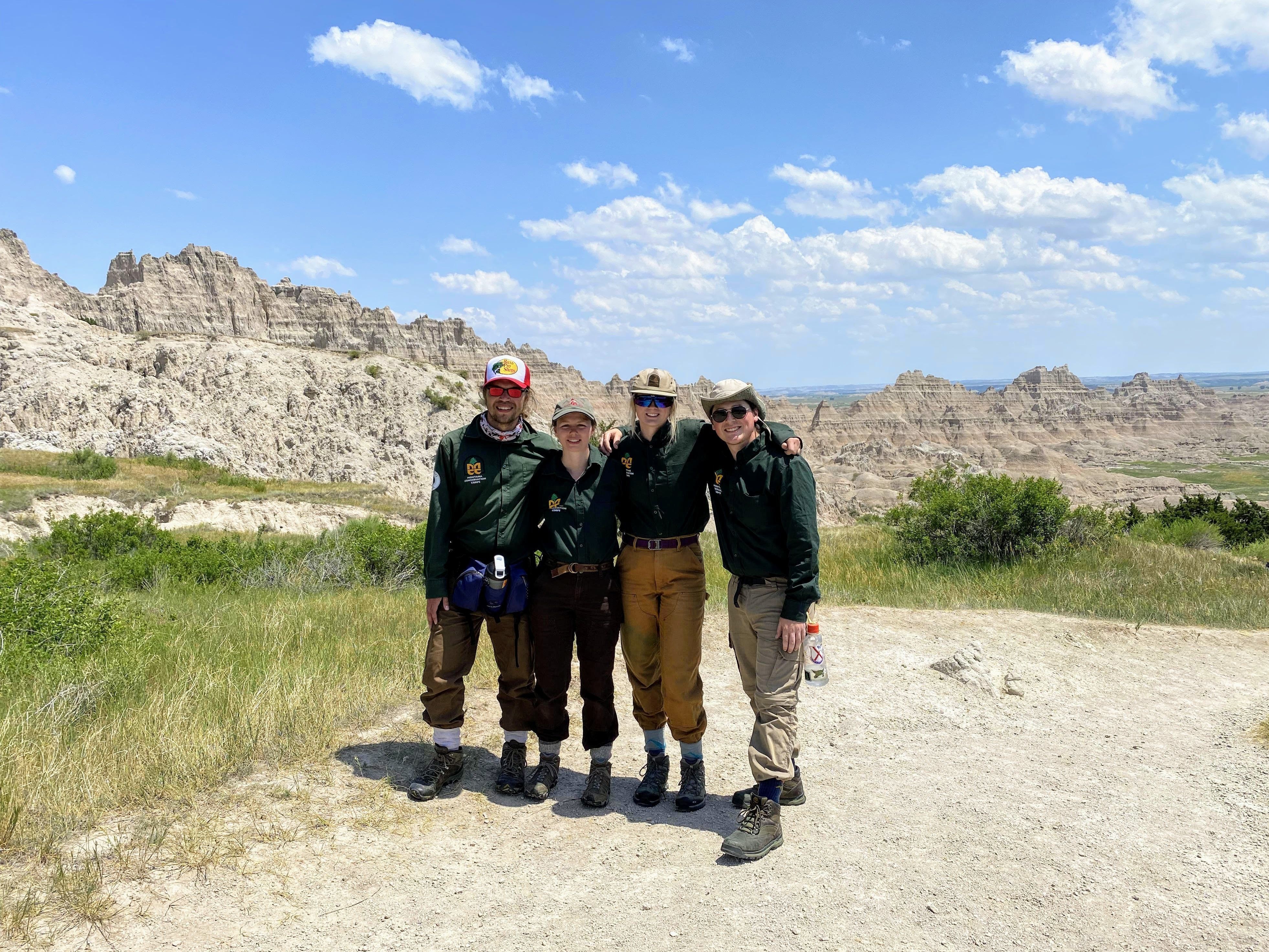 [Image description: Four MCC Members stand with their arms around one another facing the photographer, smiling. Behind them, granulated jagged peaks line the horizon.]