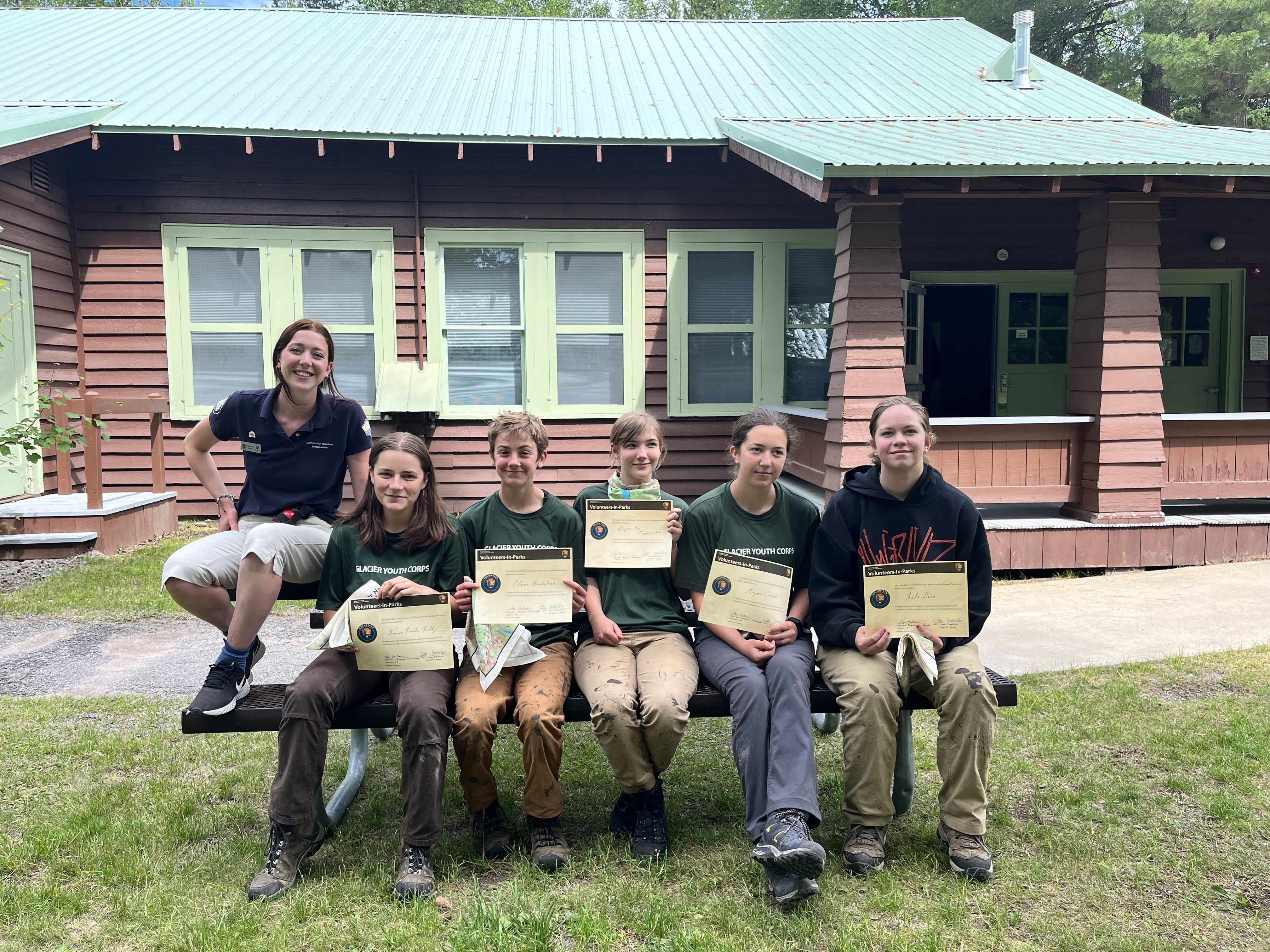 A youth crew and a leader sit smiling on a picnic table. The youth crew mates are holding certificates of completion up towards the camera.