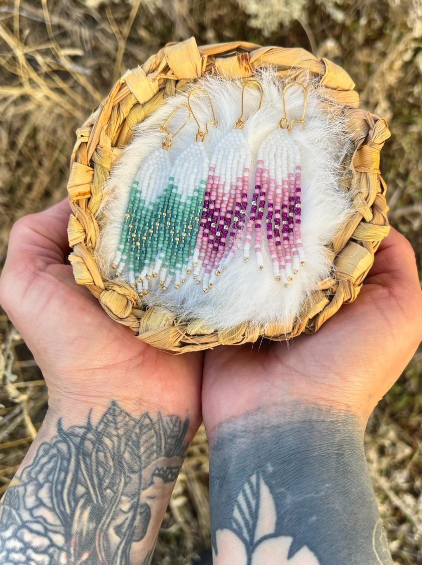 2 pairs of fringed earrings- one white and teal, the other white and pink, sit in a handmade basket held by the artist's hands. The earrings are beaded with chevron patterns. 
