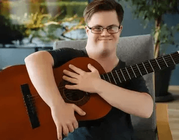 The image shows a smiling person with glasses sitting on a chair and holding an acoustic guitar. The background includes a large screen displaying a nature scene with trees and water, along with indoor plants in the room.