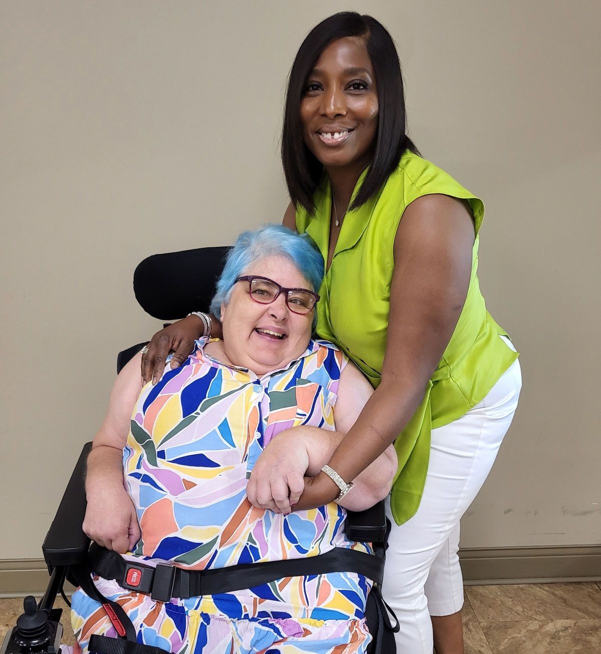 A woman with blue hair and a flower-print dress is in a wheelchair and smiles at the camera while a woman in a green top and white pants stands behind her smiling toward the camera
