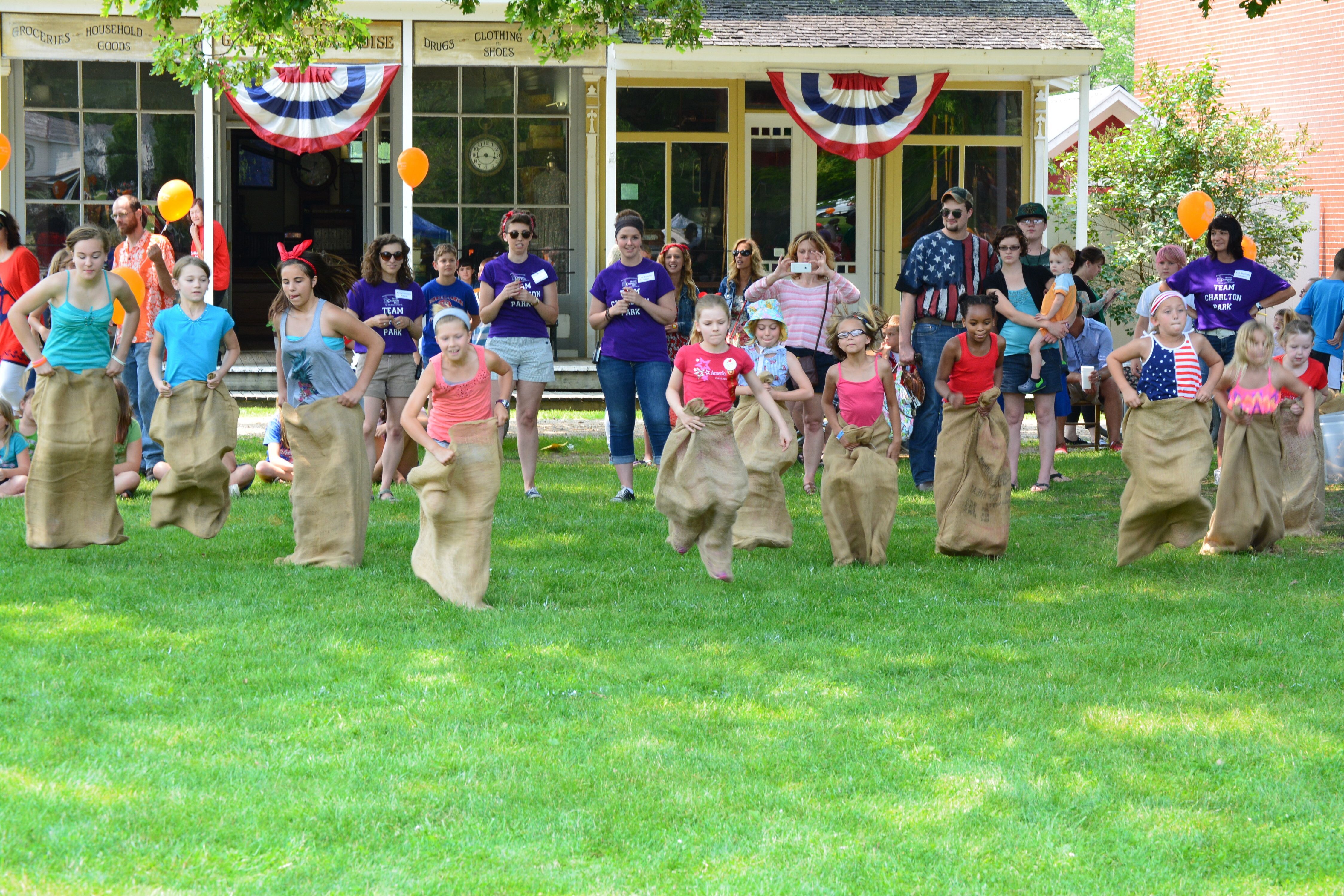Children running a sack race in coffee bean sacks on a green field with people and buildings surrounding them. 