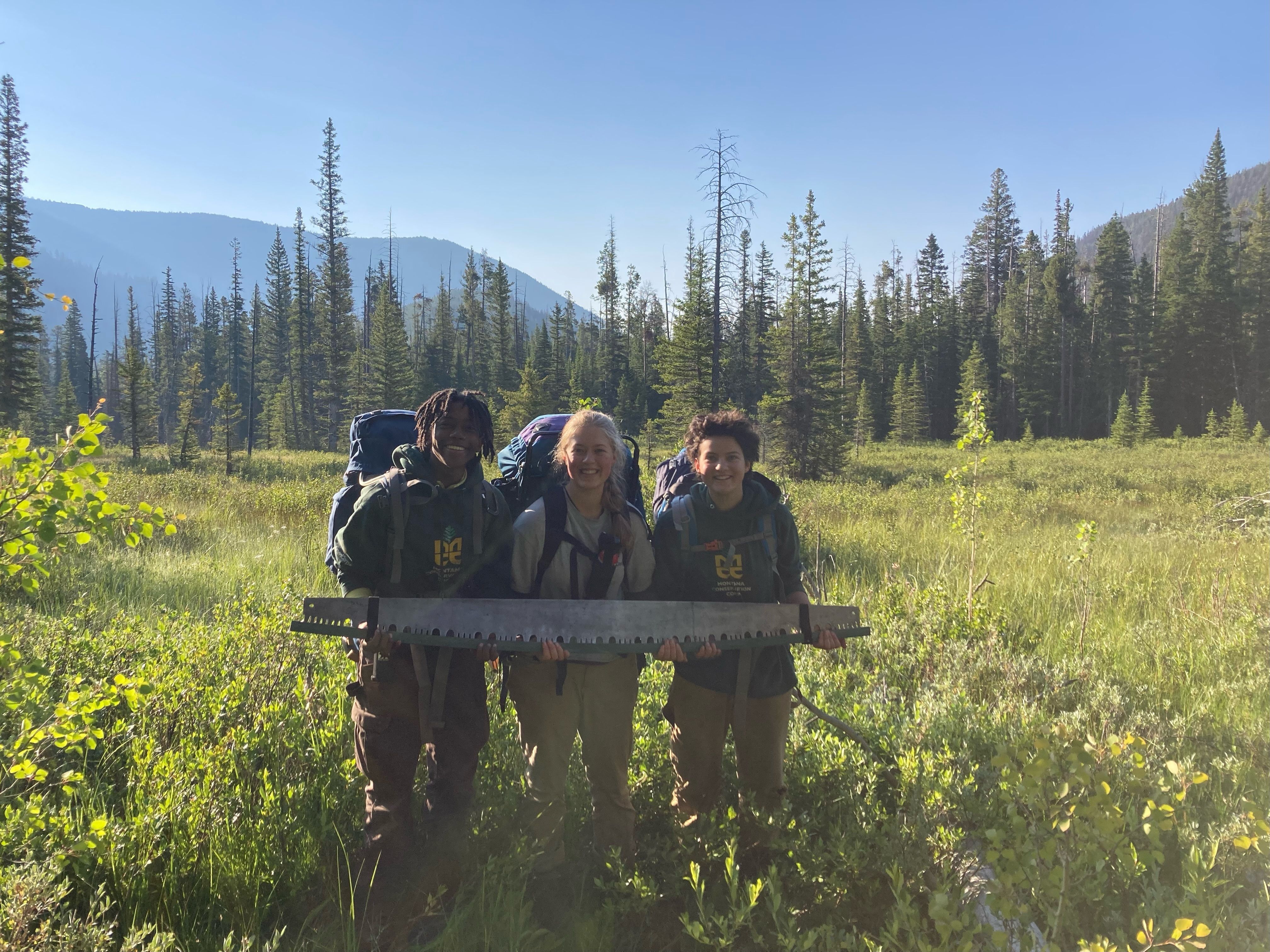 Three crew members stand smiling, holding a crosscut saw horizontally