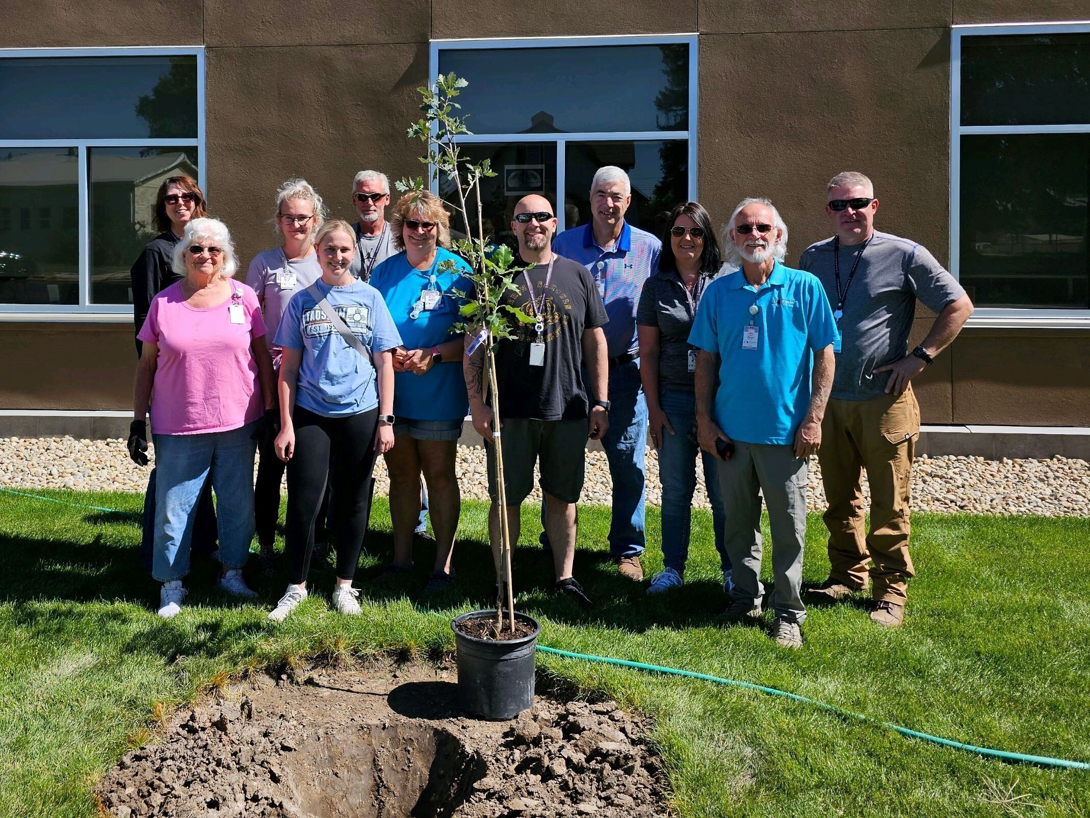 A group of people stand around a recently planted oak tree in front of an elementary school