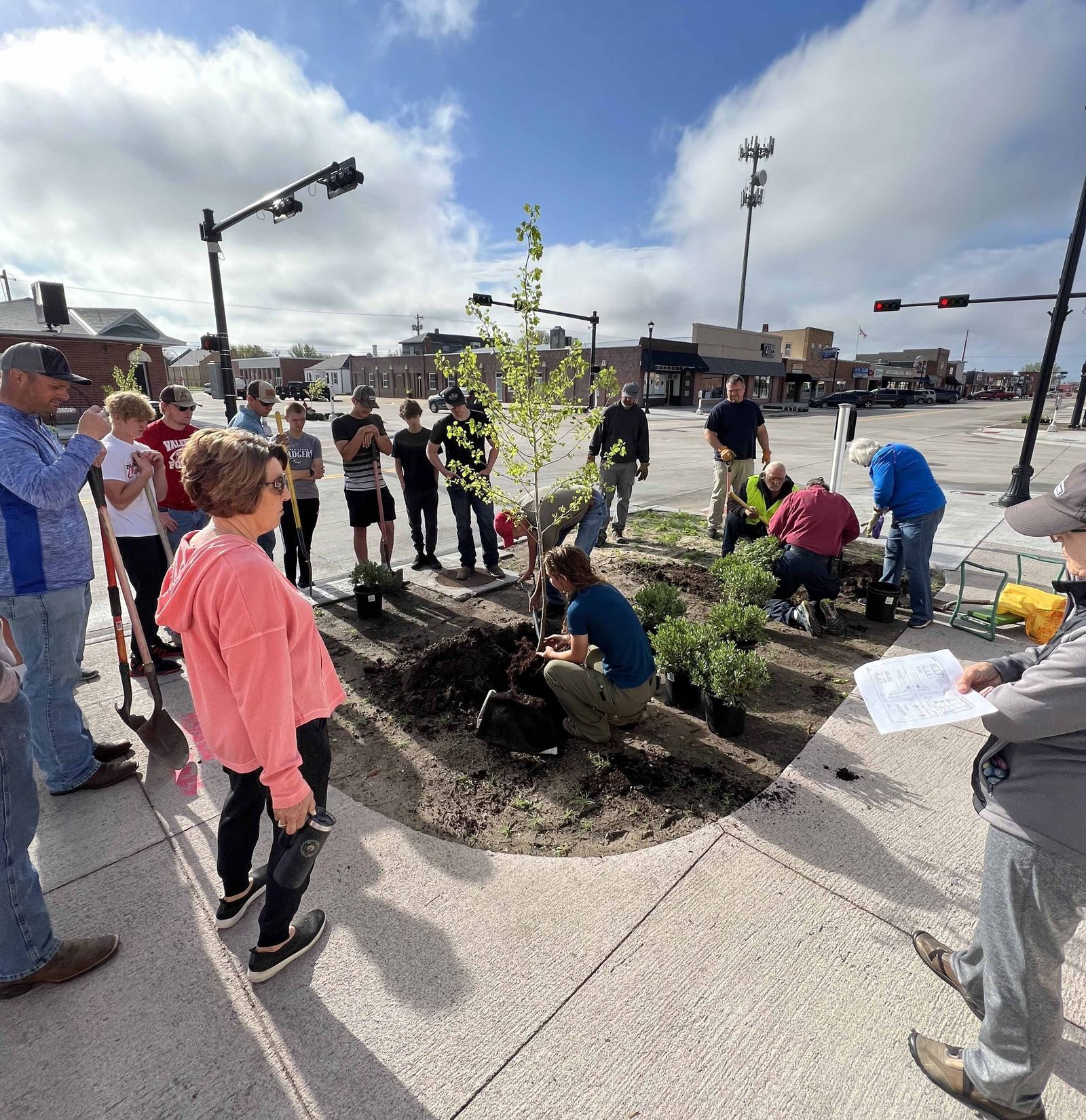 A group of high schools gather on the sidewalk around a woman who is showing them how to plant a tree. 