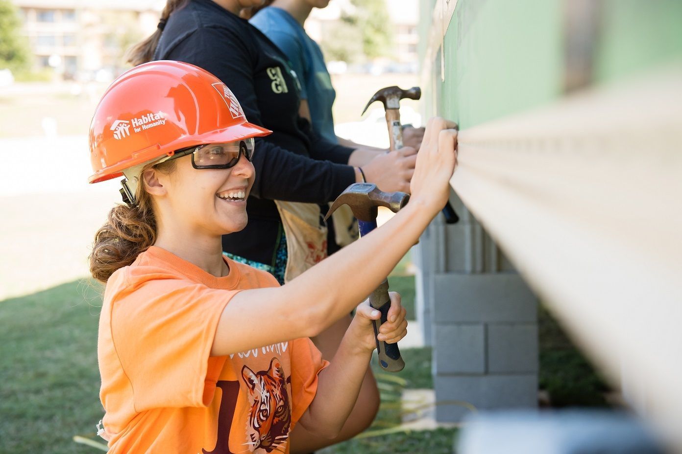 Clemson student wears an orange hard hat and hammers a nail into siding on the Homecoming house. 