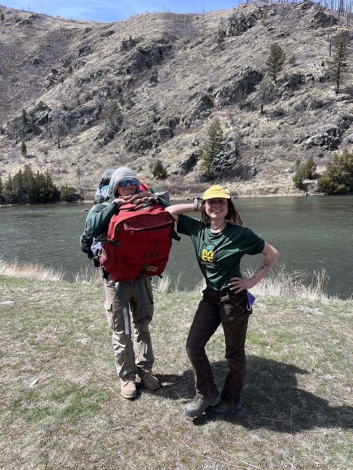 Two crew leaders stand on and bank of a river, facing the camera. One is holding a large backpack first aid kit around his front.