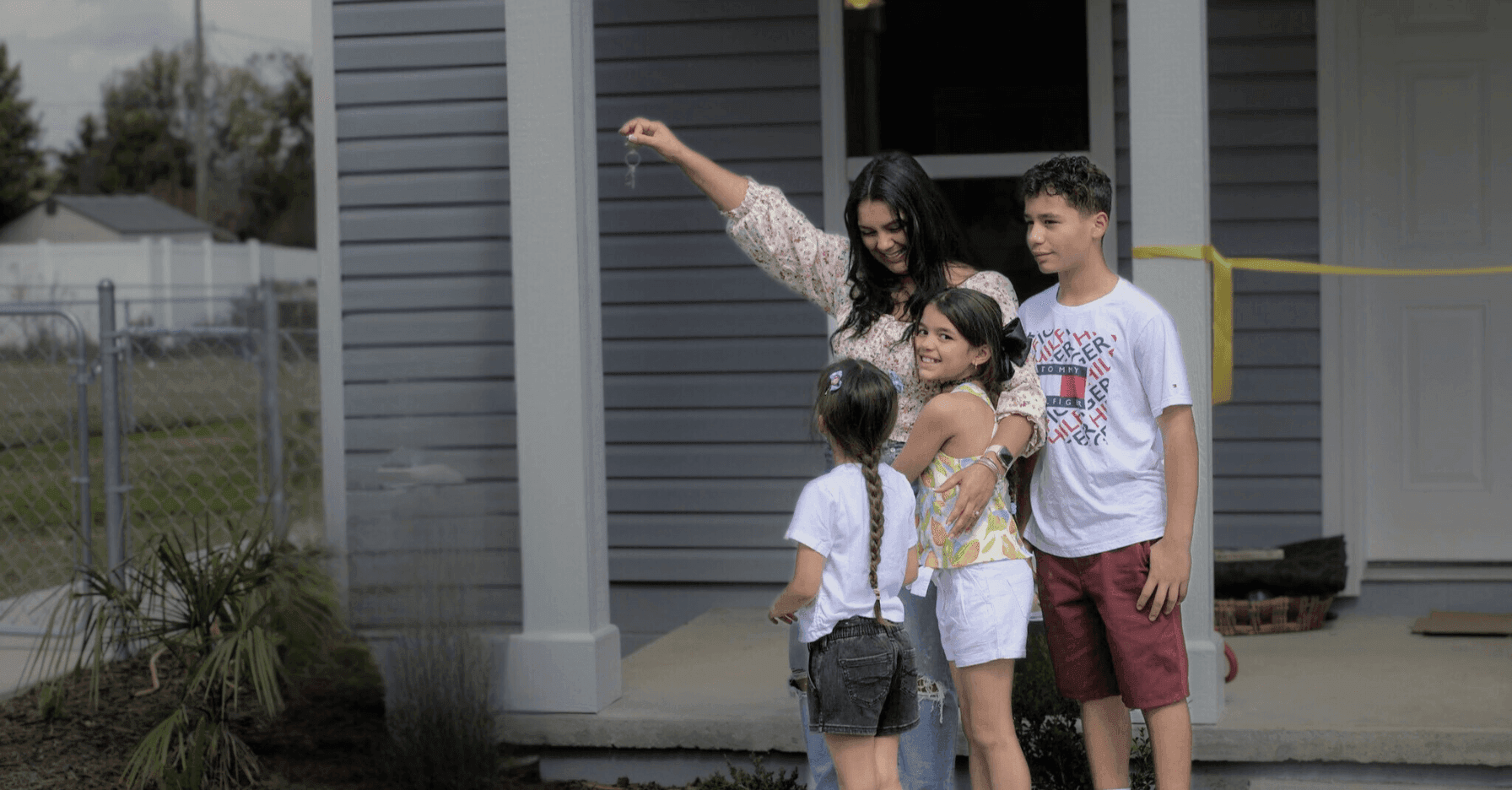 Two smiling female Habitat for Humanity volunteers in blue hard hats and shirts pose in front of a home under construction. One woman raises her fist in a cheerful, empowered gesture.