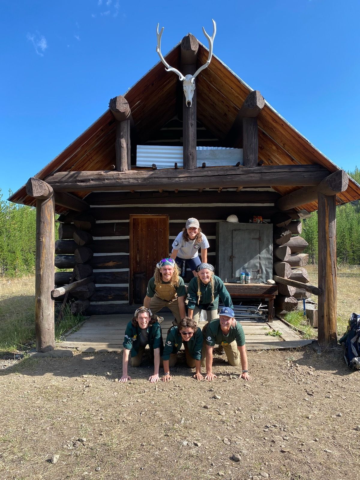 A crew forms a human pyramid. Behind them is a NPS cabin.