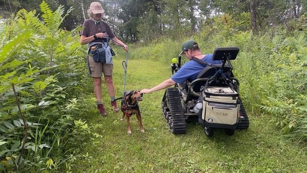 an owner with their dog stopping on a walking trail for a participant in a wheelchair to give pets to their dog. 