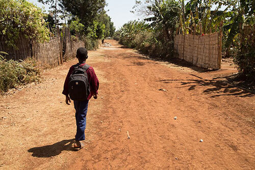 A young boy with a backpack walks down a dusty road lined with vegetation on a sunny day.