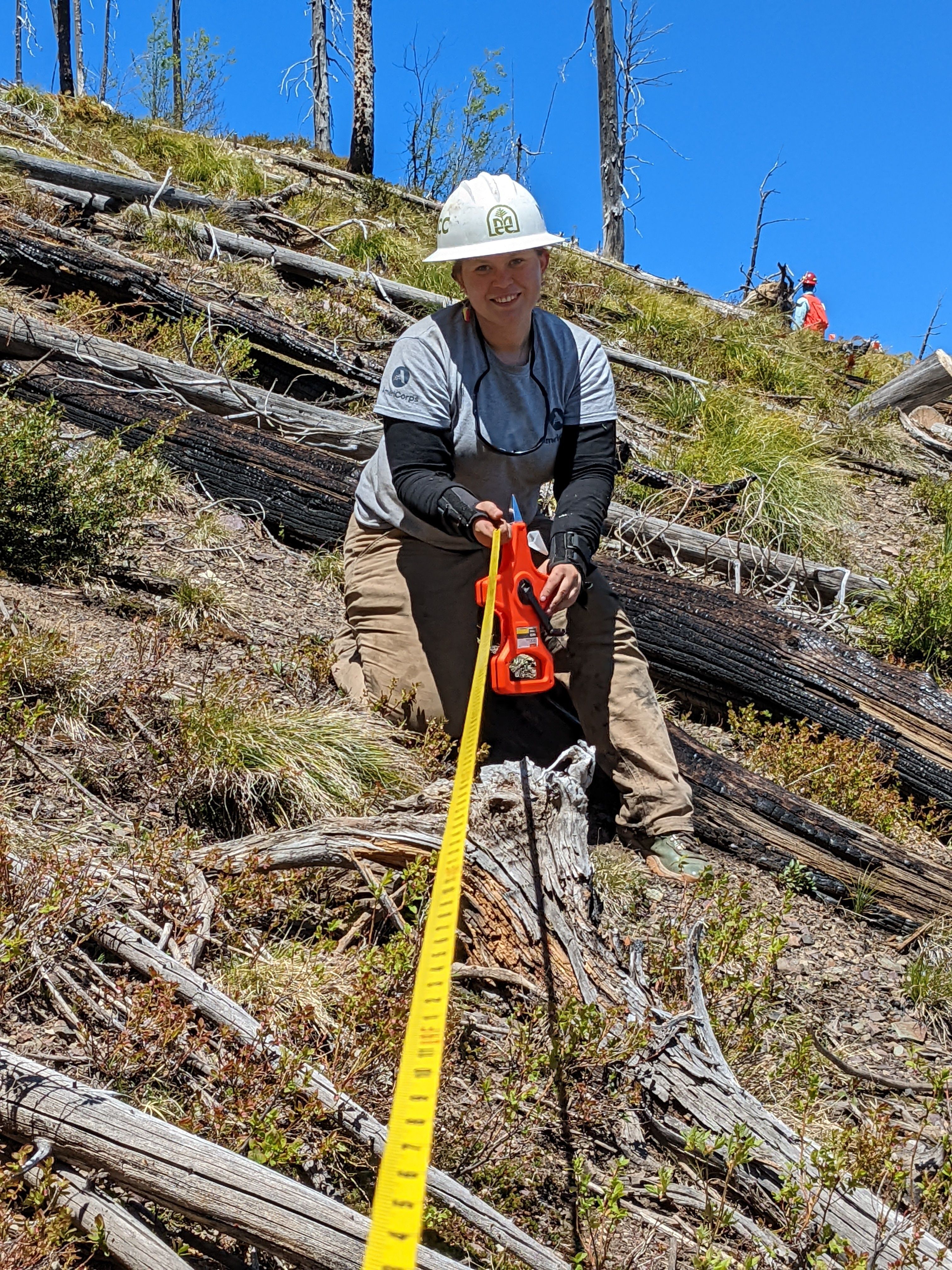 A crew leader kneels uphill from the photographer. They are holding a long tape measure that the photographer is holding onto the other end of.