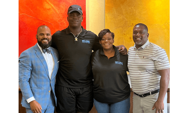 Photo of Marcus Williams and NIL Emery Jones, Jones' mother Nikki, and Jones' former athletic coach Pat Green. The group are standing very close together, smiling, and celebrating against a red ochre and yellow ochre background.