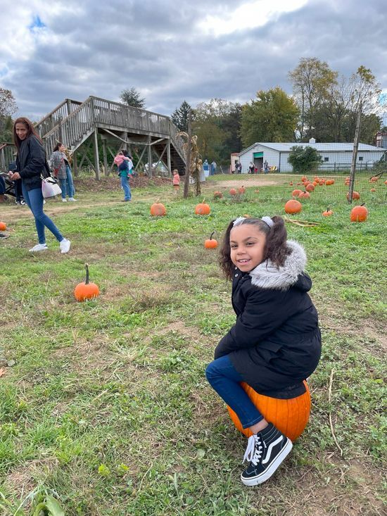 A young girl with a big smile sits on a pumpkin she's picked at Snipes Farm Fall Festival