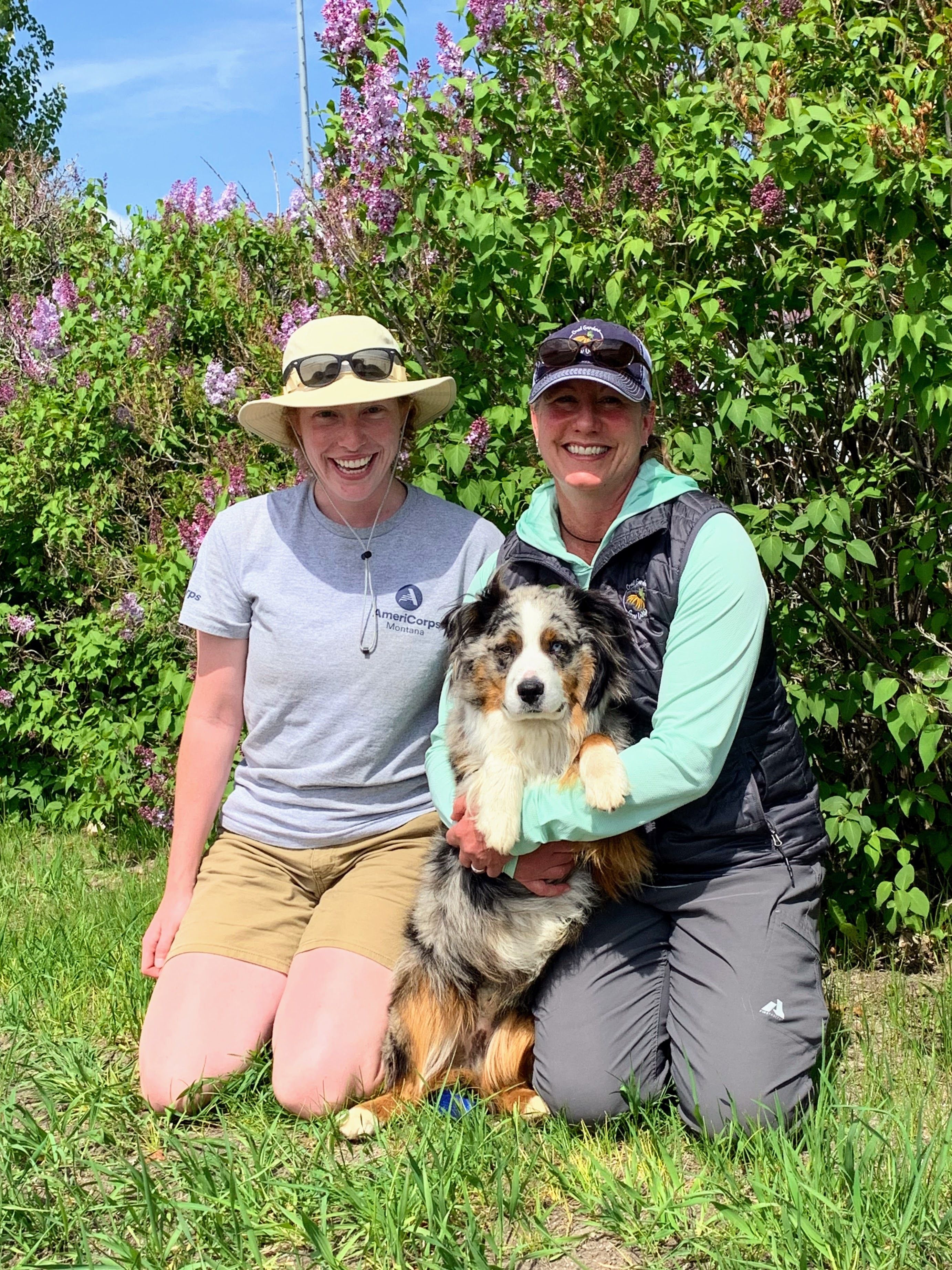 Two women sit in the grass, smiling at the camera. One is holding a dog.