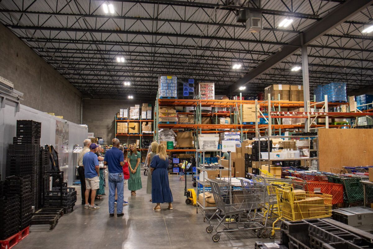 Warehouse interior with people gathered, shelves stacked with boxes, and shopping carts lined up.