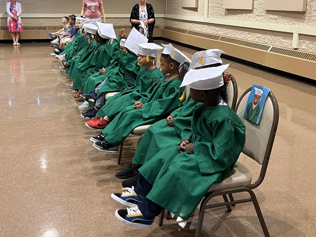 Students sit in while caps and green gowns in a row of chairs for their preschool graduation.