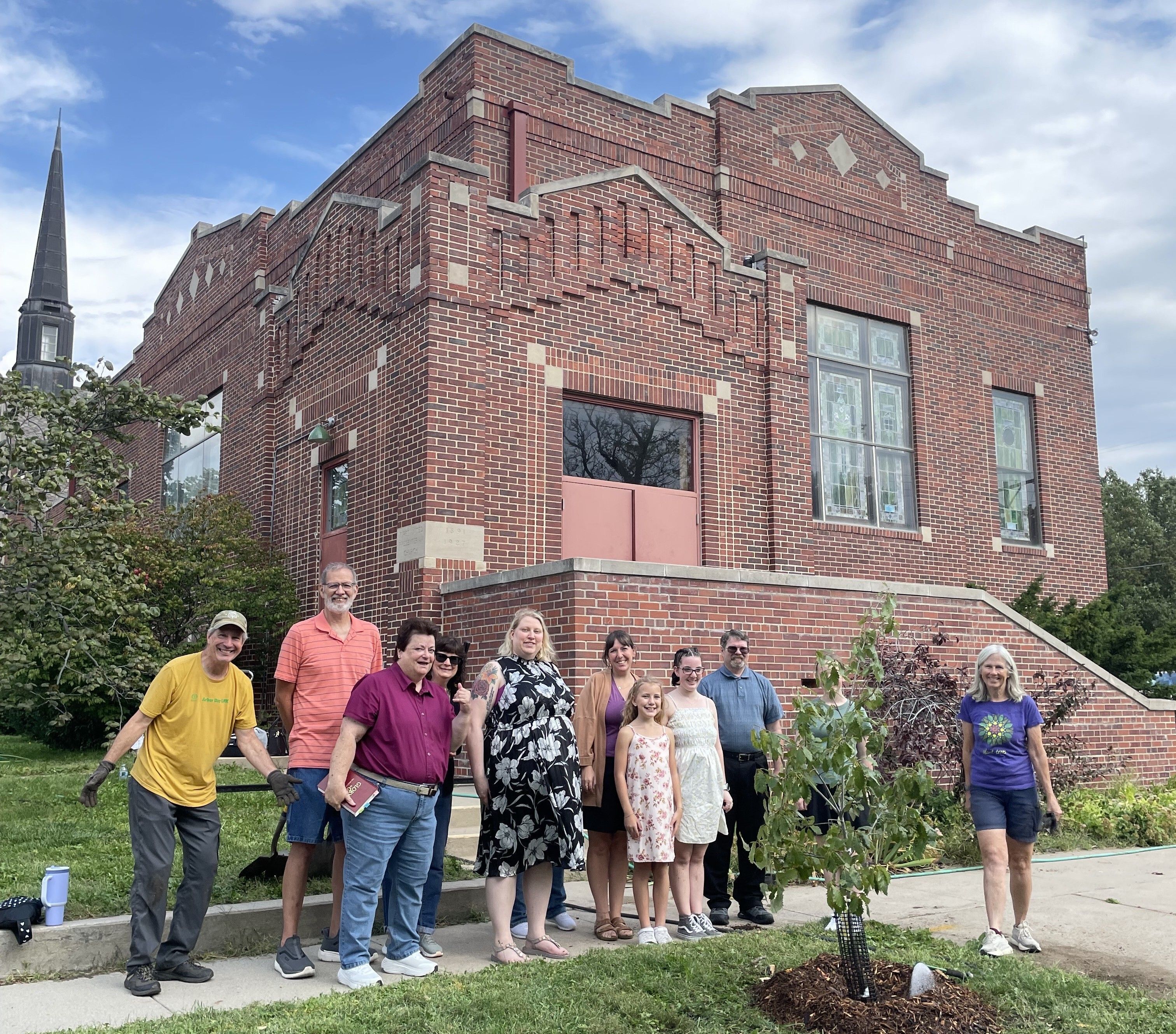 A group of people stand next to a newly planted tree. 