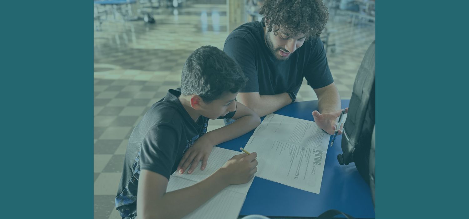 A young boy is studying, and a young man is instructing him.