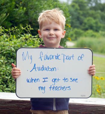 Young boy holds a white board with writing that reads "My favorite part of Audubon: when I get to see my teachers"