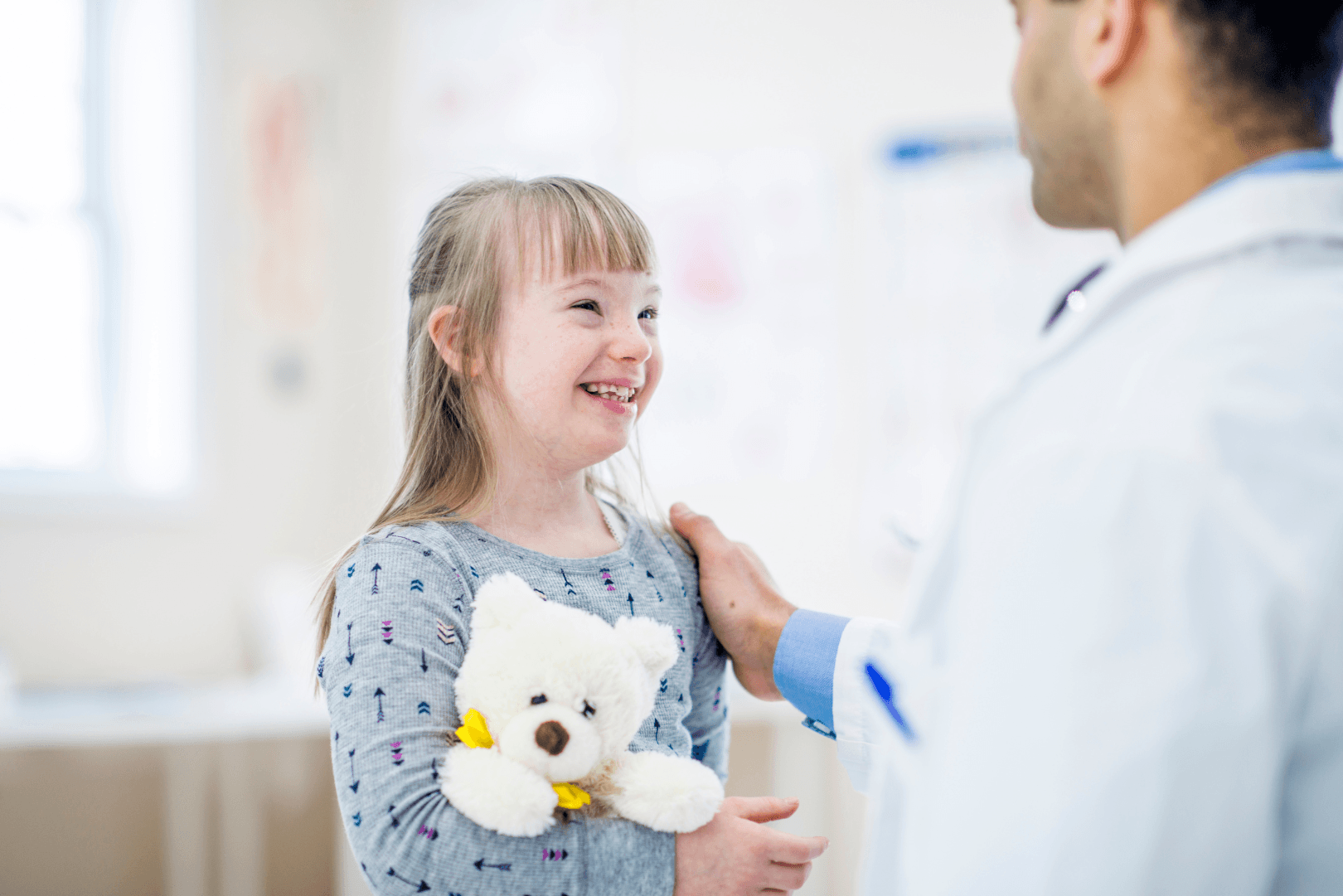 a young girl with light brown hair holds a teddy bear at an annual appointment with her pediatrician 