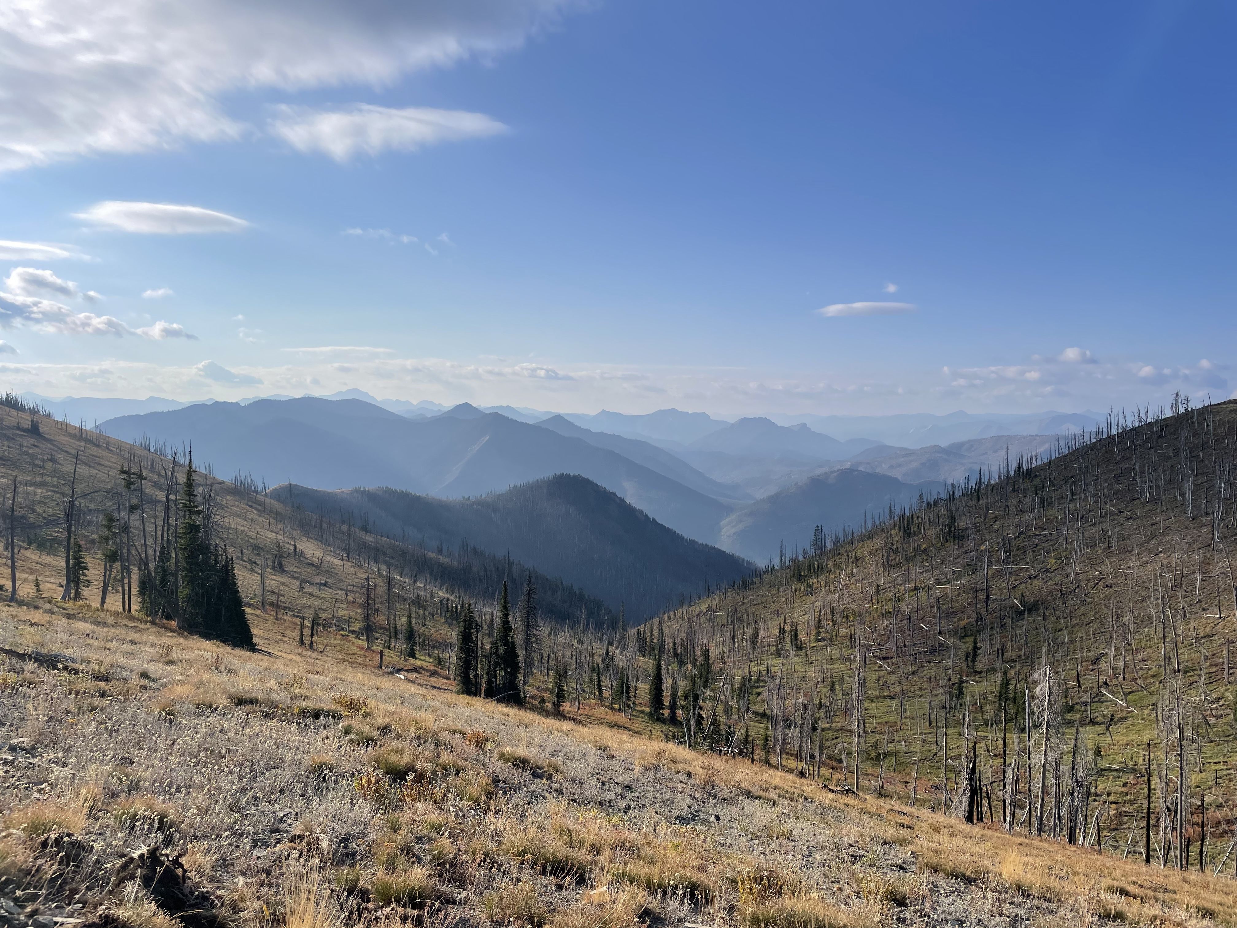 A view from a hillside down into a mountain valley. The peaks in the distance are blue and hazy, backed by blue sky.