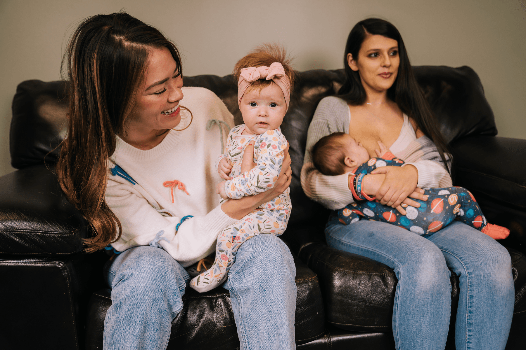 Two women, sitting on a brown leather sofa. The woman on the left is asian and smiling at her baby on her lap. Her baby is female, wearing white and pink pajamas, and has a pink headband bow in her hair. The mom on the right is Latina and is breastfeeding