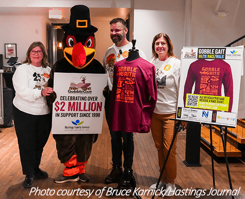 Group posing with Mercy the turkey who is holding a sign stating over two million dollars raised in support of Hastings Family Service, next to 2024 race shirt on display.