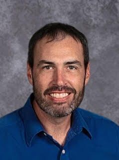 Professional headshot of a smiling middle-aged man with short dark hair, salt-and-pepper beard and brown eyes. He wears a vibrant blue button-up shirt and poses against a textured gray backdrop.