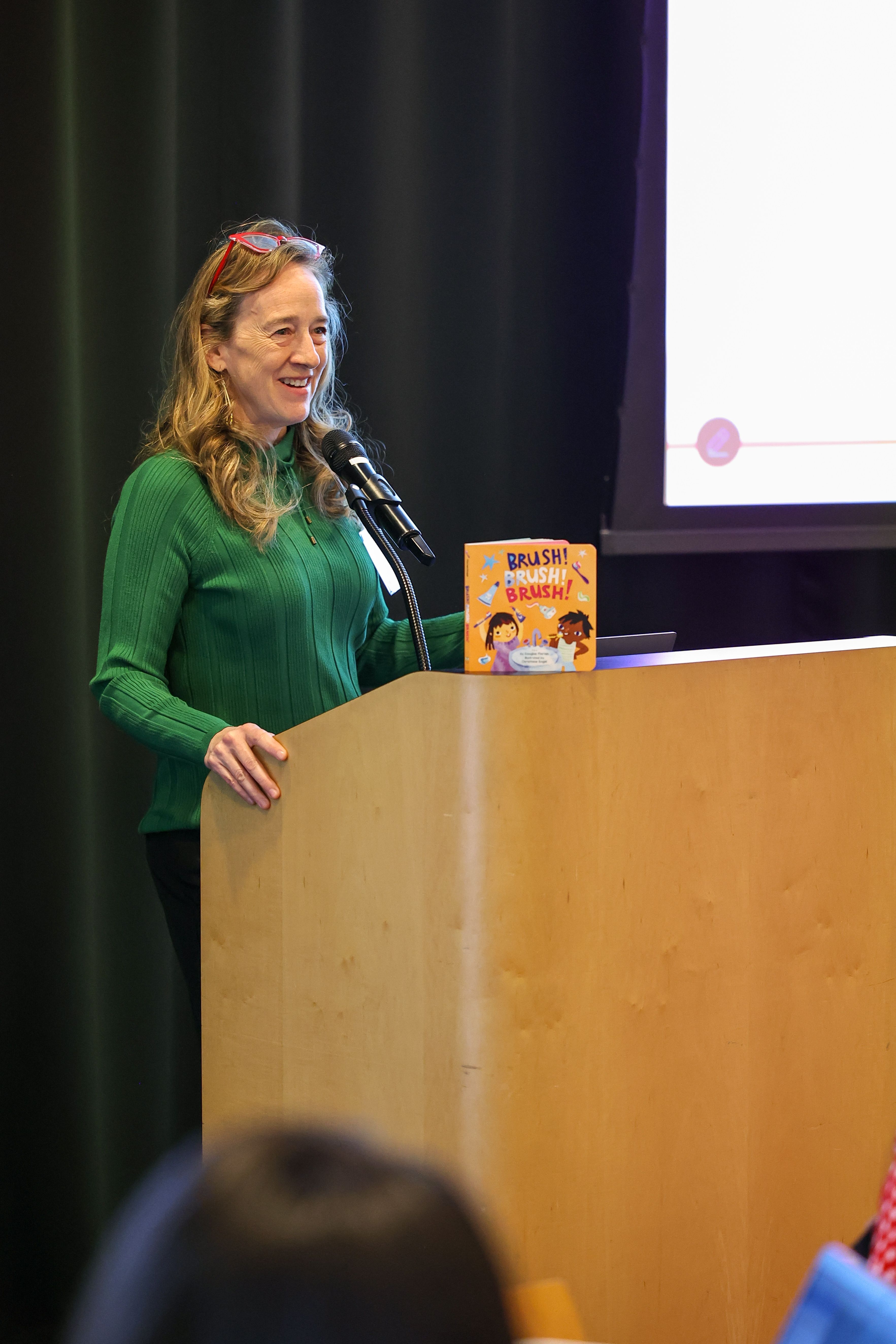 Woman with long hair and green sweater stands at podium with children's book