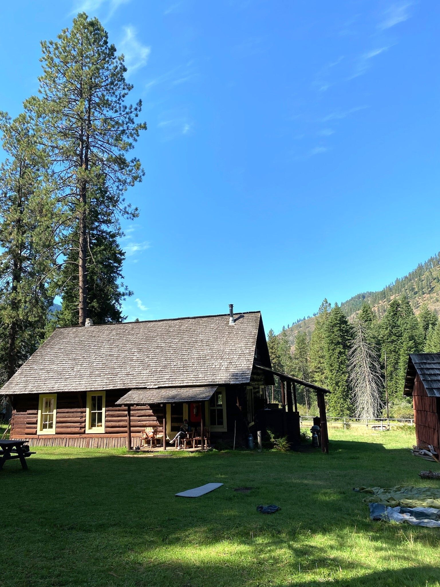 A cabin in a field with tall trees behind and on a hill side in the distance.