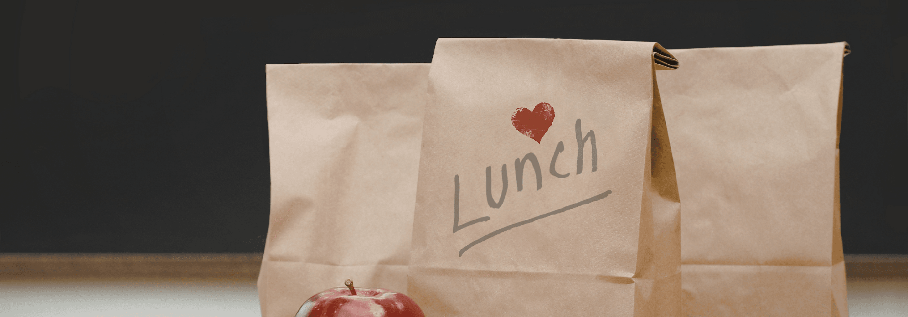 Three brown bagged lunches and a red apple on a wood surface with a black background. One lunch bag has "lunch" written on it in marker with a drawn red heart above it.