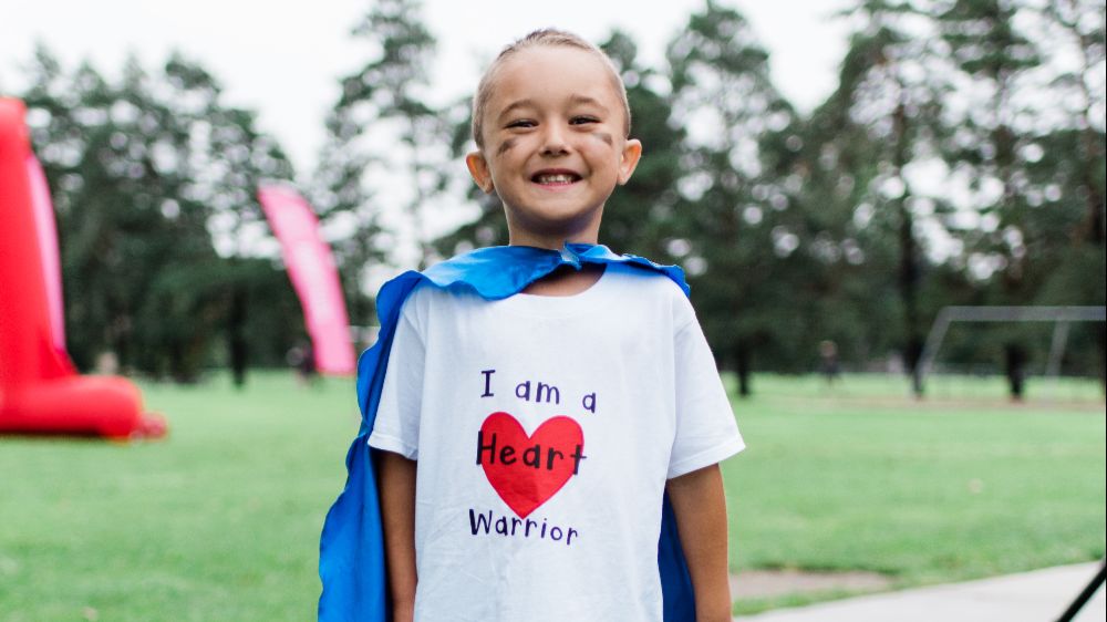 Young child smiling, wearing a blue cape and white t-shirt with the words "I am a Heart Warrior" on the front.