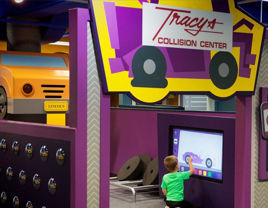 A child interacts with a touchscreen at "Tracy's Collision Center," a play area with a car theme.